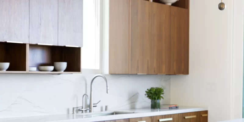 Transitional-style kitchen featuring a sleek quartzite waterfall island with subtle veining. The kitchen design includes walnut cabinetry, white quartzite countertops, a stainless steel faucet, and contemporary brass lighting fixtures. A decorative bowl of artichokes adds a natural touch to the clean, minimal aesthetic.