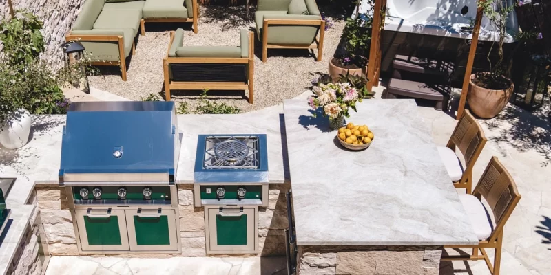 An outdoor kitchen with a stone countertop featuring built-in stainless steel barbecue and stovetop grill. The setup includes seating with two chairs, a bowl of lemons, and a vase of flowers on the countertop. The background includes a seating area with green cushioned outdoor furniture and a hot tub, surrounded by plants and gravel landscaping.